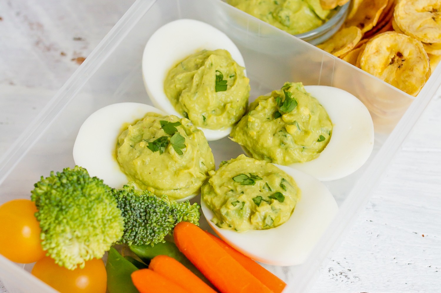 Avocado deviled eggs in a meal prep container. Each hard-boiled egg half features a guacamole inspired filling. Served alongside fresh vegetables and homemade plantain chips. | peteandgerrys.com 