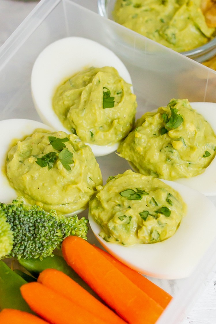 Avocado deviled eggs in a meal prep container. Each hard-boiled egg half features a guacamole inspired filling. Served alongside fresh vegetables and homemade plantain chips. | peteandgerrys.com 