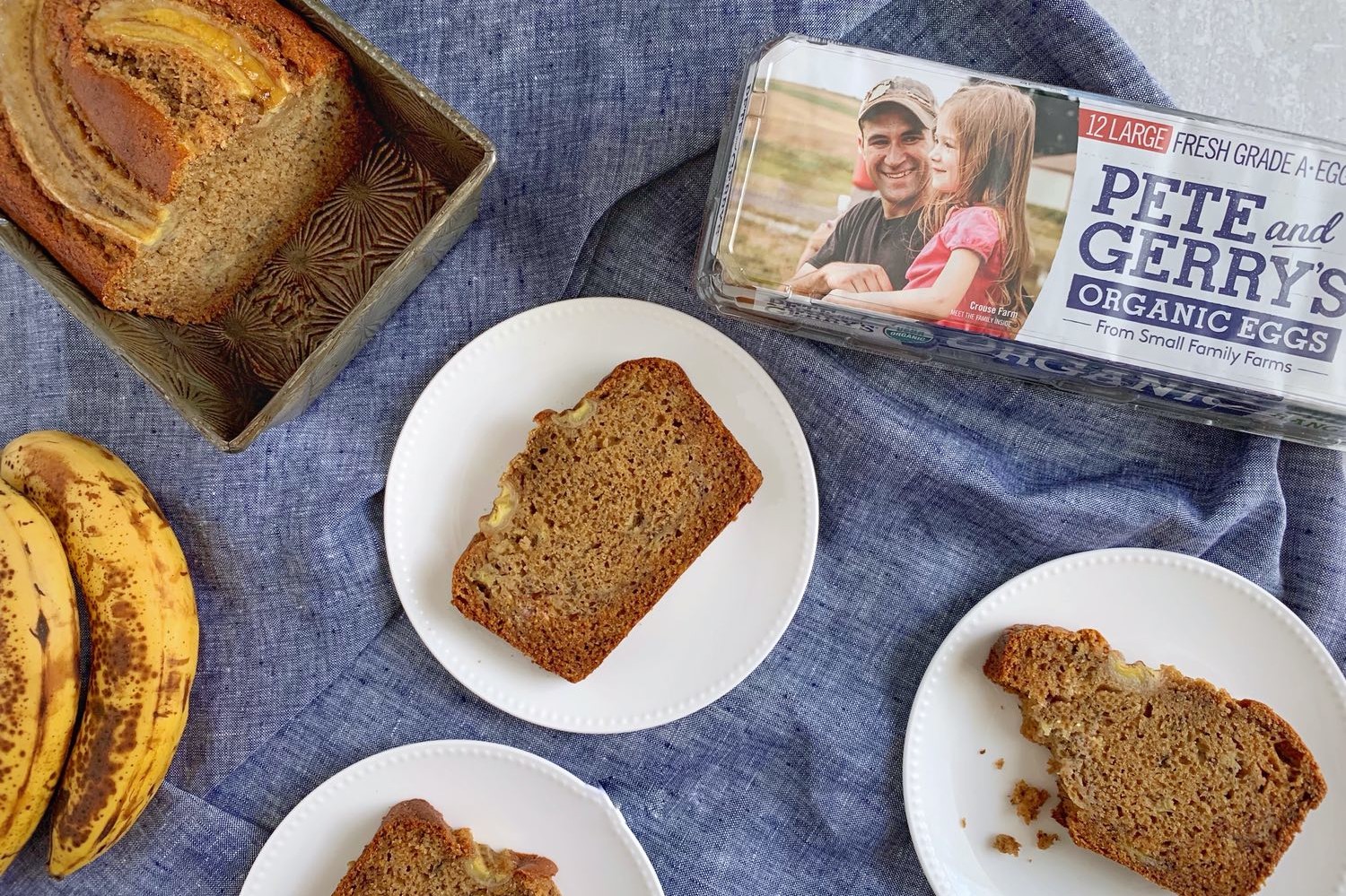 Brown butter banana bread with banana baked in top for decoration. Slices are plated on white dishes and show a moist crumb. Pete and Gerry's Organic Egg carton in the corner of the frame. | peteandgerrys.com 