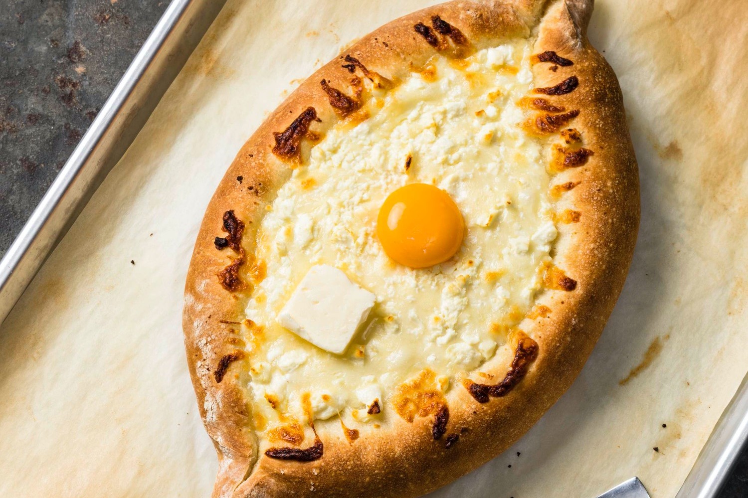 Adjaruli khachapuri (often called a Georgian cheese bread boat) is a marriage of yeasted bread, butter, egg yolk, feta, and mozzarella cheese. Shown placed on a baking sheet for serving.