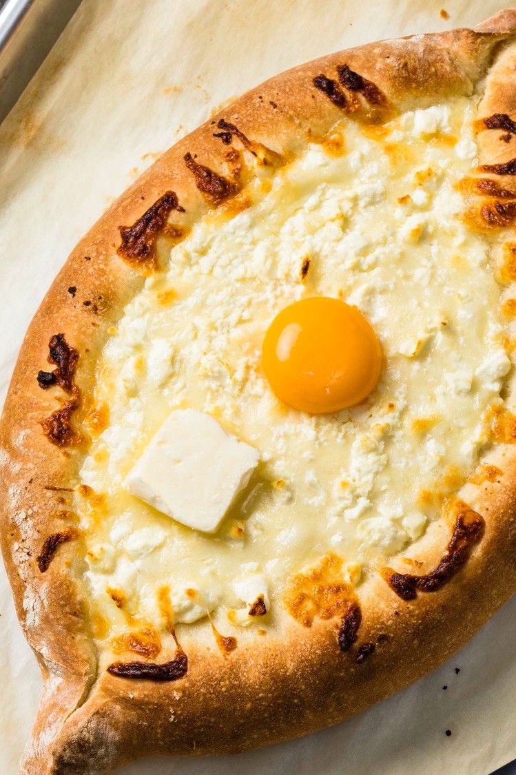 Adjaruli khachapuri (often called a Georgian cheese bread boat) is a marriage of yeasted bread, butter, egg yolk, feta, and mozzarella cheese. Shown placed on a baking sheet for serving.