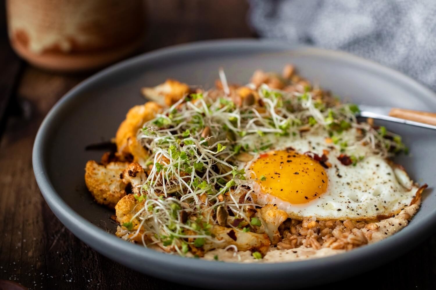 A nutritious vegetarian grain bowl for lunch or dinner. Bowl features roasted cauliflower florets, hummus, farro, microgreens, and a fried organic egg. | peteandgerrys.com 