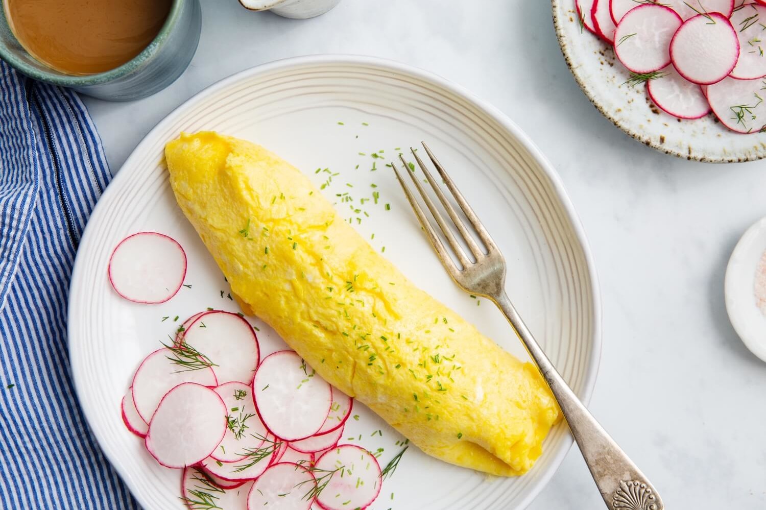 A classic French omelette made with organic eggs and butter, boasting a golden yellow exterior and soft, custardy interior. The omelette is topped with minced chives and served with a side of thinly sliced radishes. | peteandgerrys.com