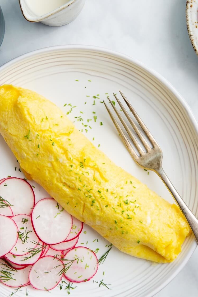 A classic French omelette made with organic eggs and butter, boasting a golden yellow exterior and soft, custardy interior. The omelette is topped with minced chives and served with a side of thinly sliced radishes. | peteandgerrys.com