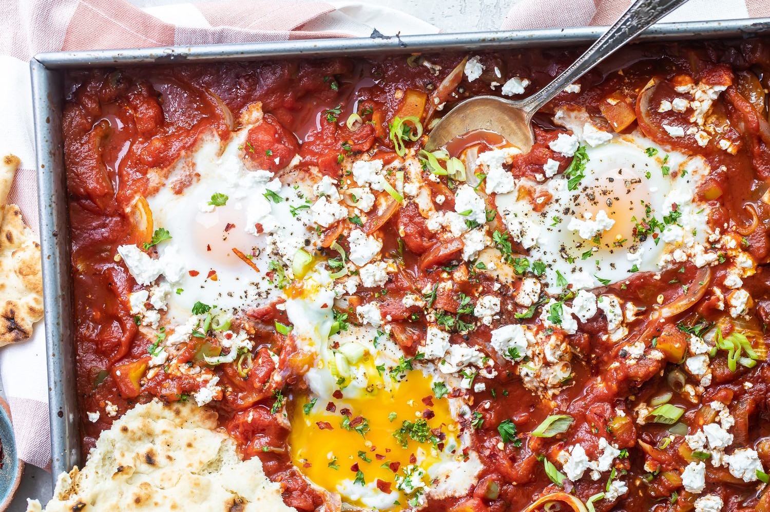 Shakshuka prepared in a sheet pan, topped with parsley and crumbled feta, and served alongside warm pita.